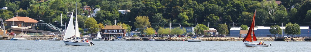 wide scene showing two sailboats, one following the other in a race. In the background is the shoreline with the boat club building.